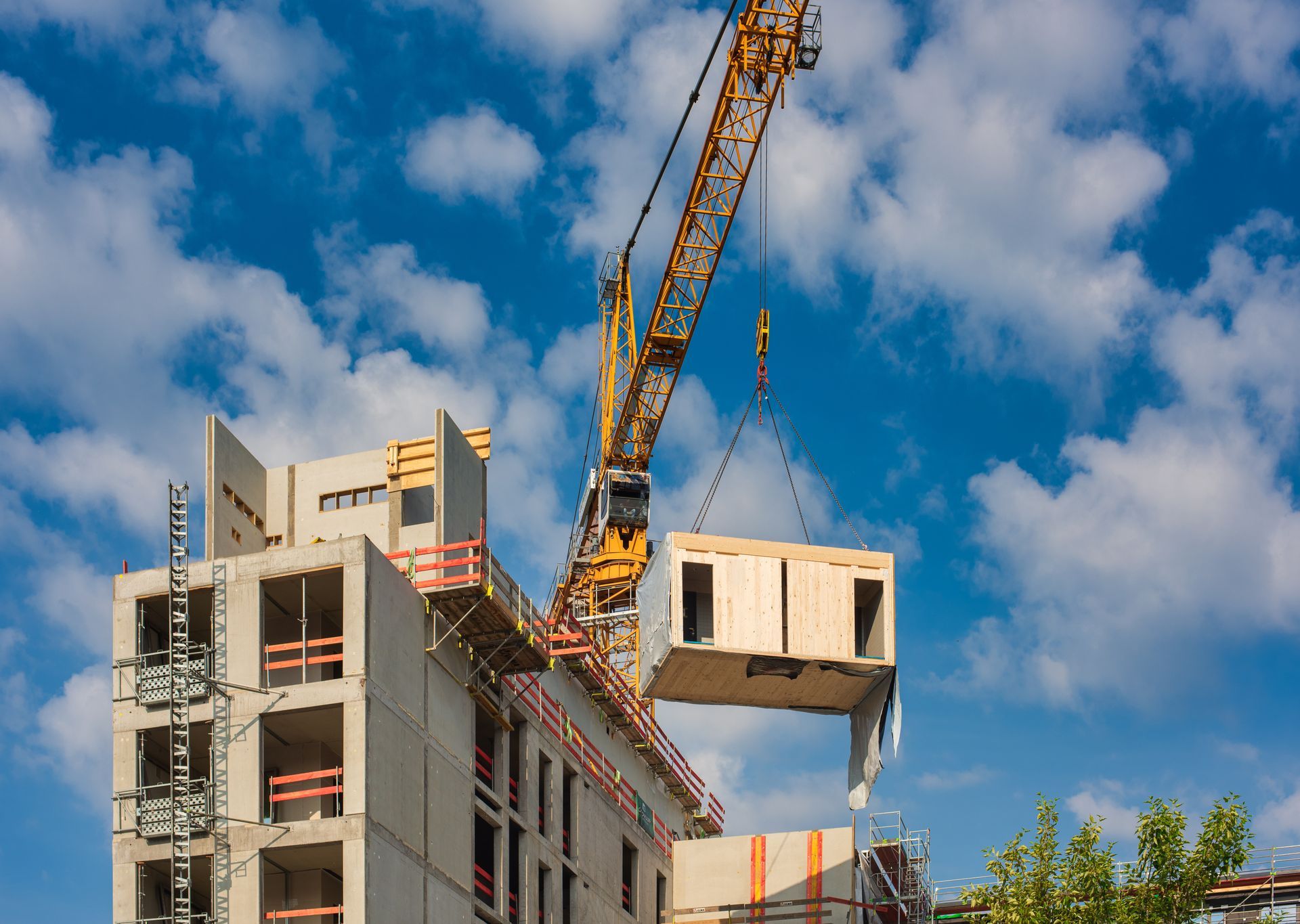 Grue soulevant un élément de construction modulaire sur une structure en béton partiellement construite, sous un ciel nuageux.