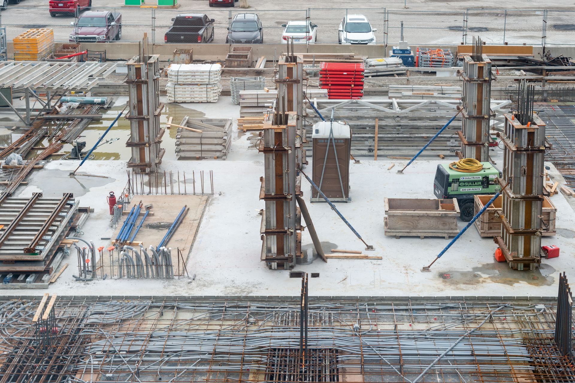 Chantier de construction avec piliers en béton, machines et matériaux ; voitures en arrière-plan.