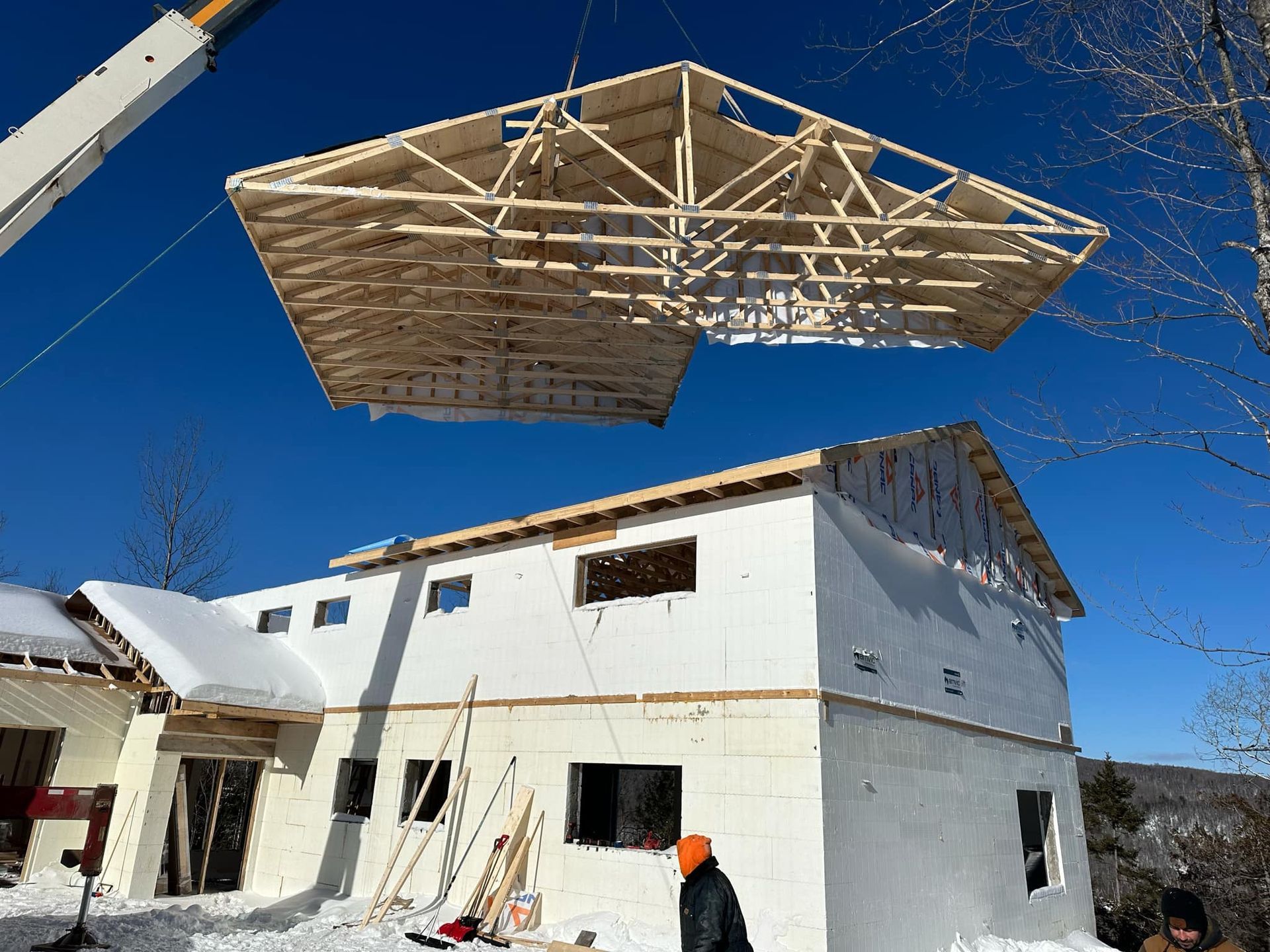 Une grue soulève une ferme de toit au-dessus d'une maison à deux étages en construction, dans un décor enneigé.