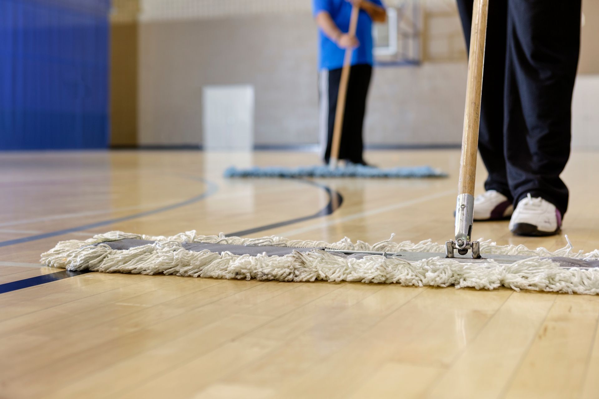 Two people are mopping a wooden floor in a gym.
