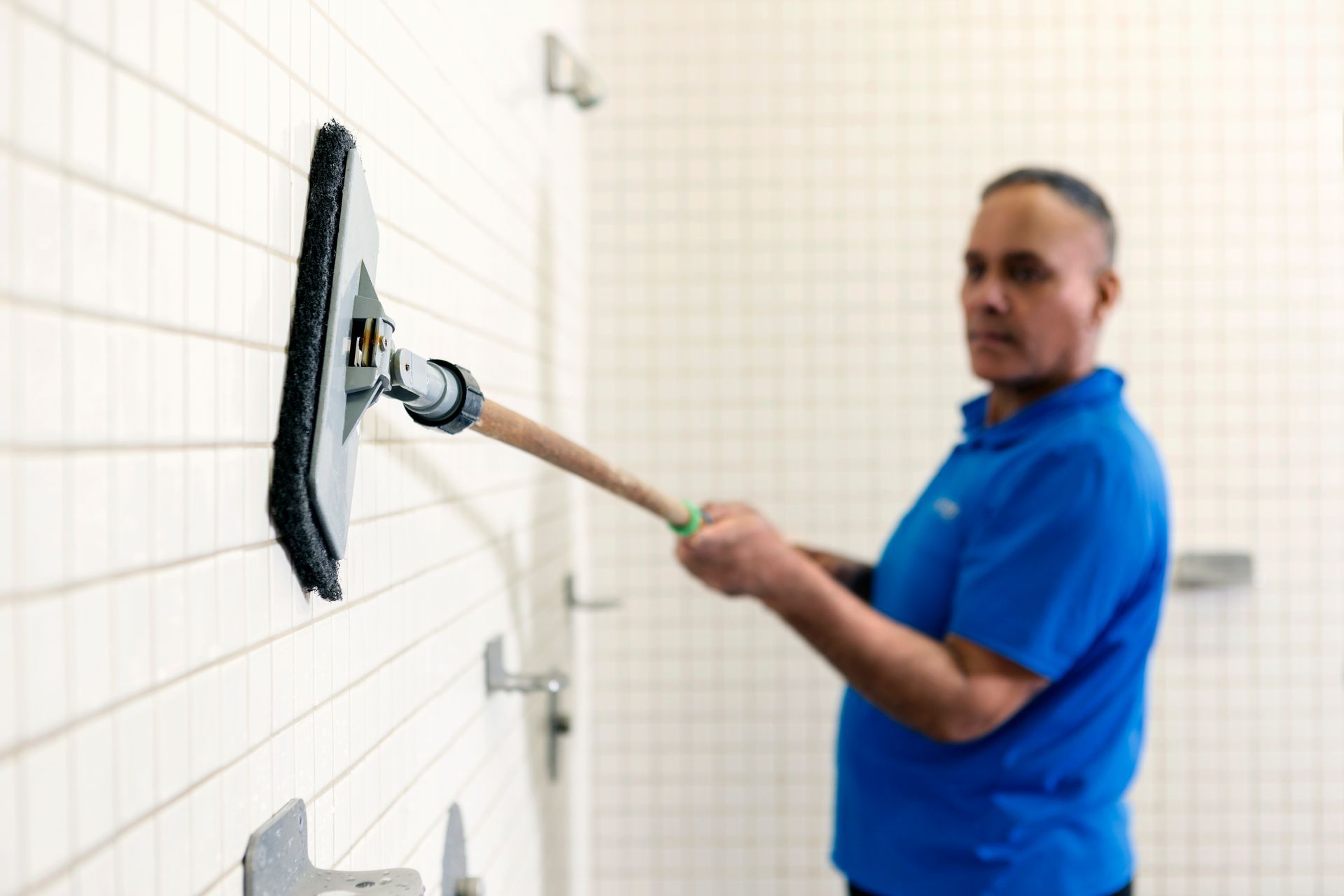 A man in a blue shirt is cleaning a bathroom wall with a mop.