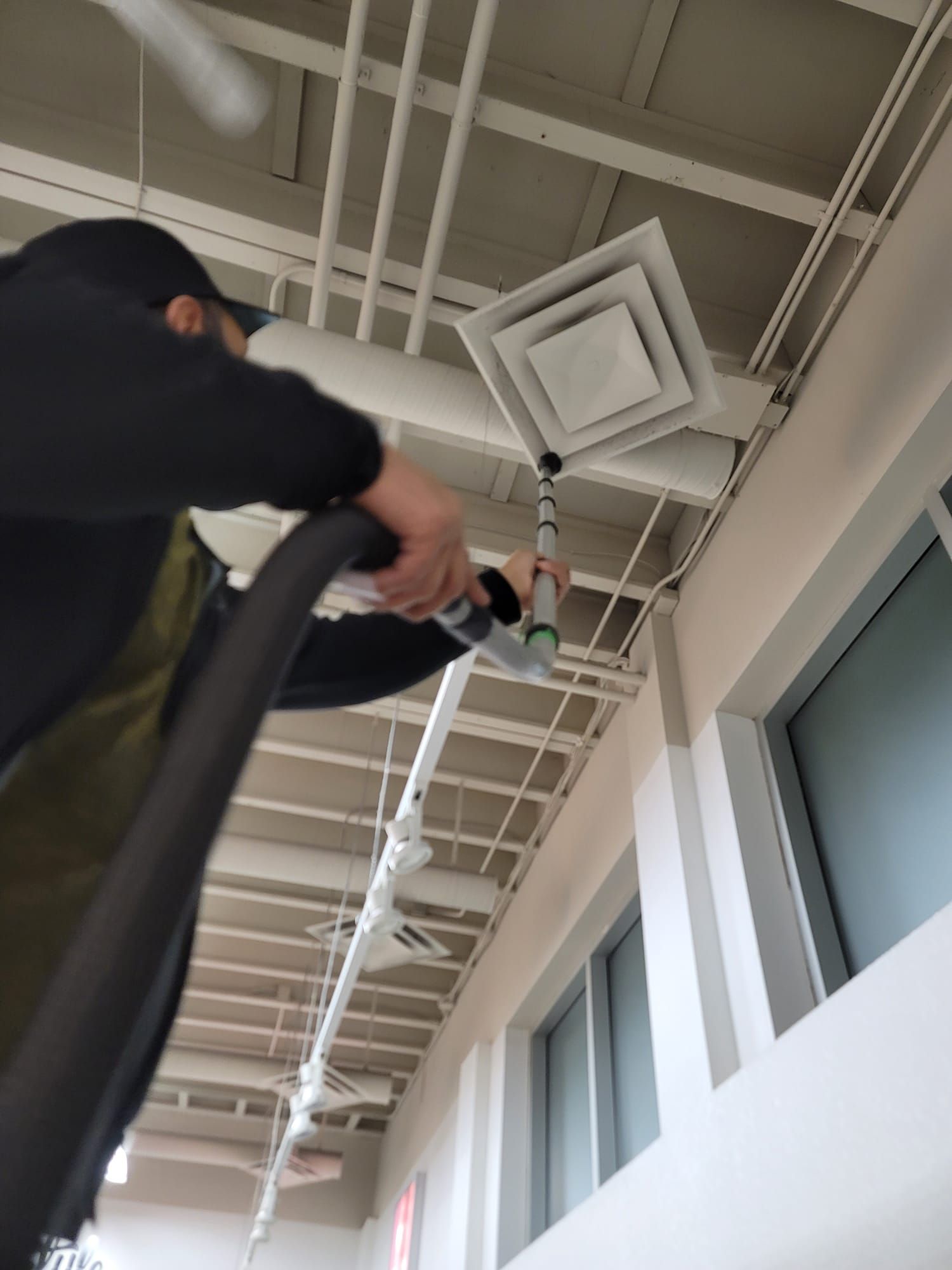 Person vacuuming a ceiling air vent in a building. They are holding the hose with a long extension.