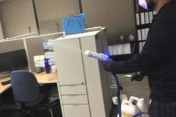 Person disinfecting an office with a spray, near a filing cabinet and desk.