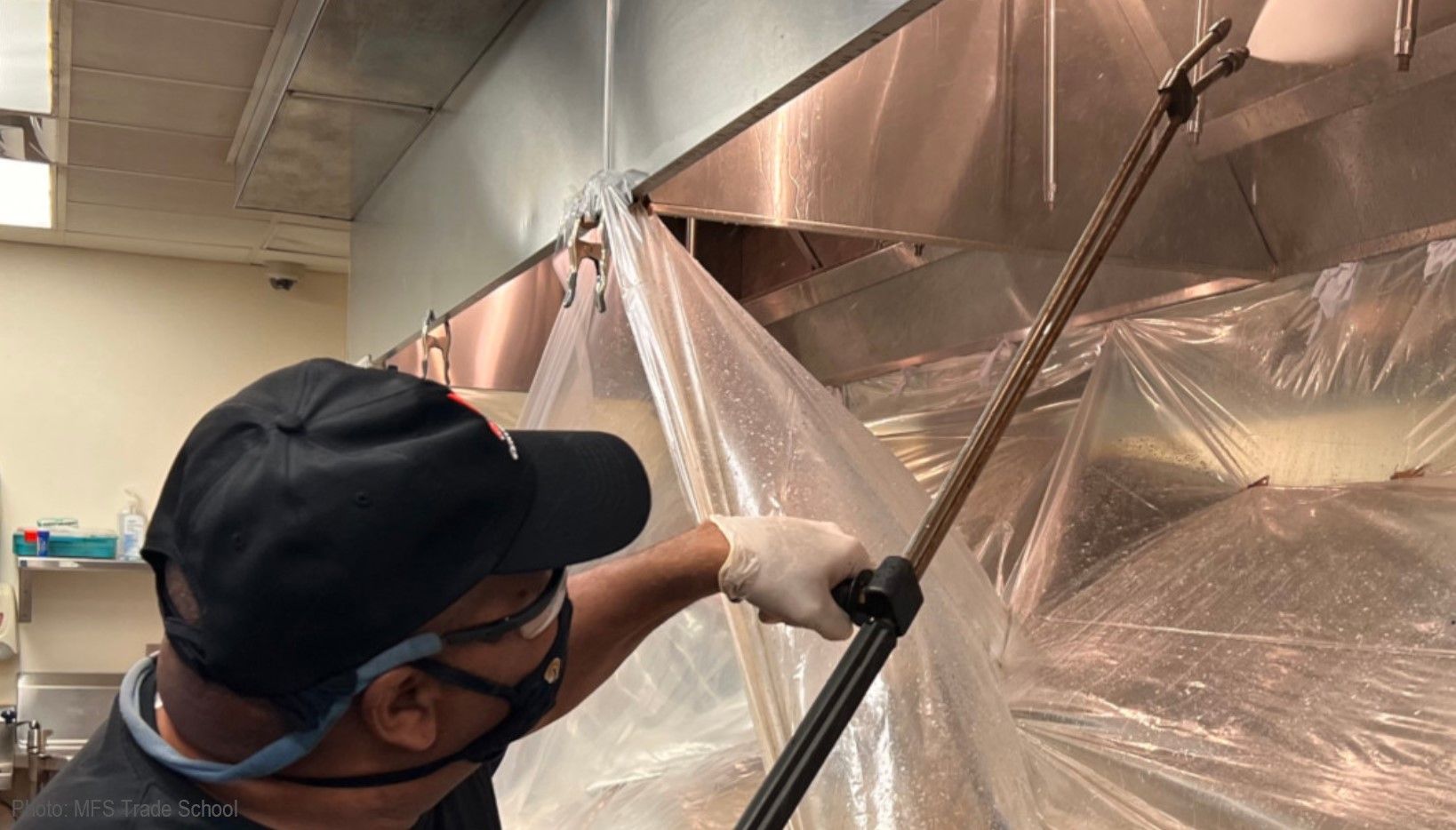 A man wearing a mask is cleaning a kitchen hood with a sprayer.