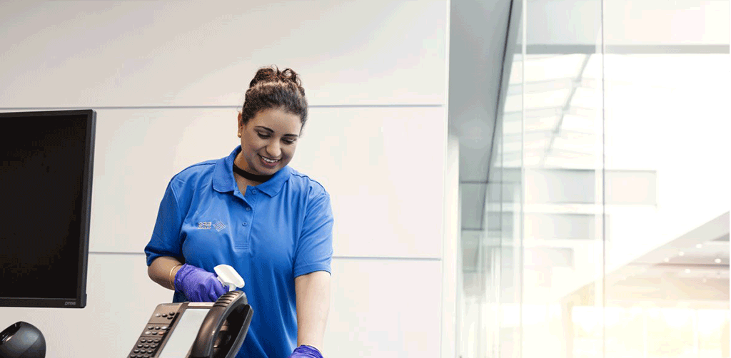 A woman in a blue shirt and gloves is cleaning a treadmill in an office.