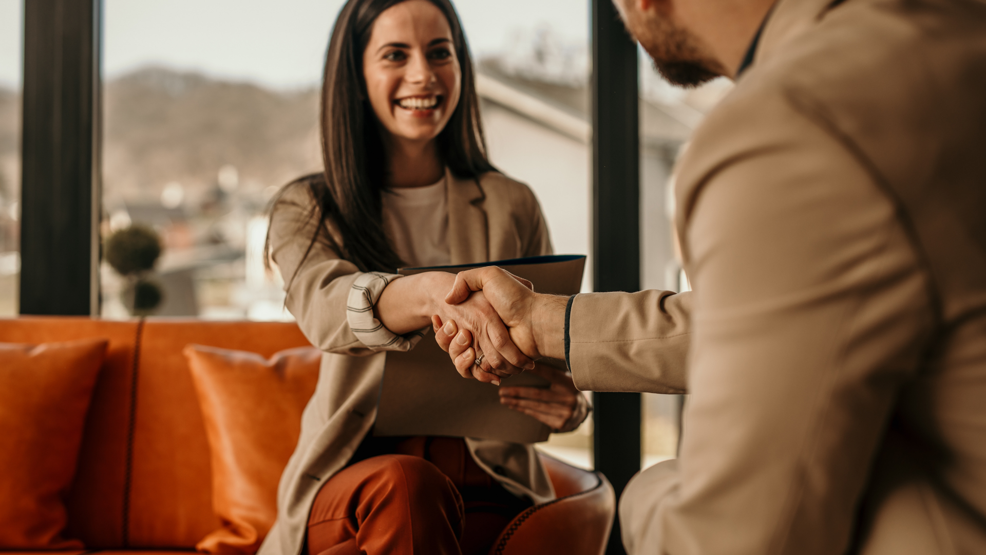 Two people shaking hands in a bright office lounge, with an orange couch and smiling woman in the background