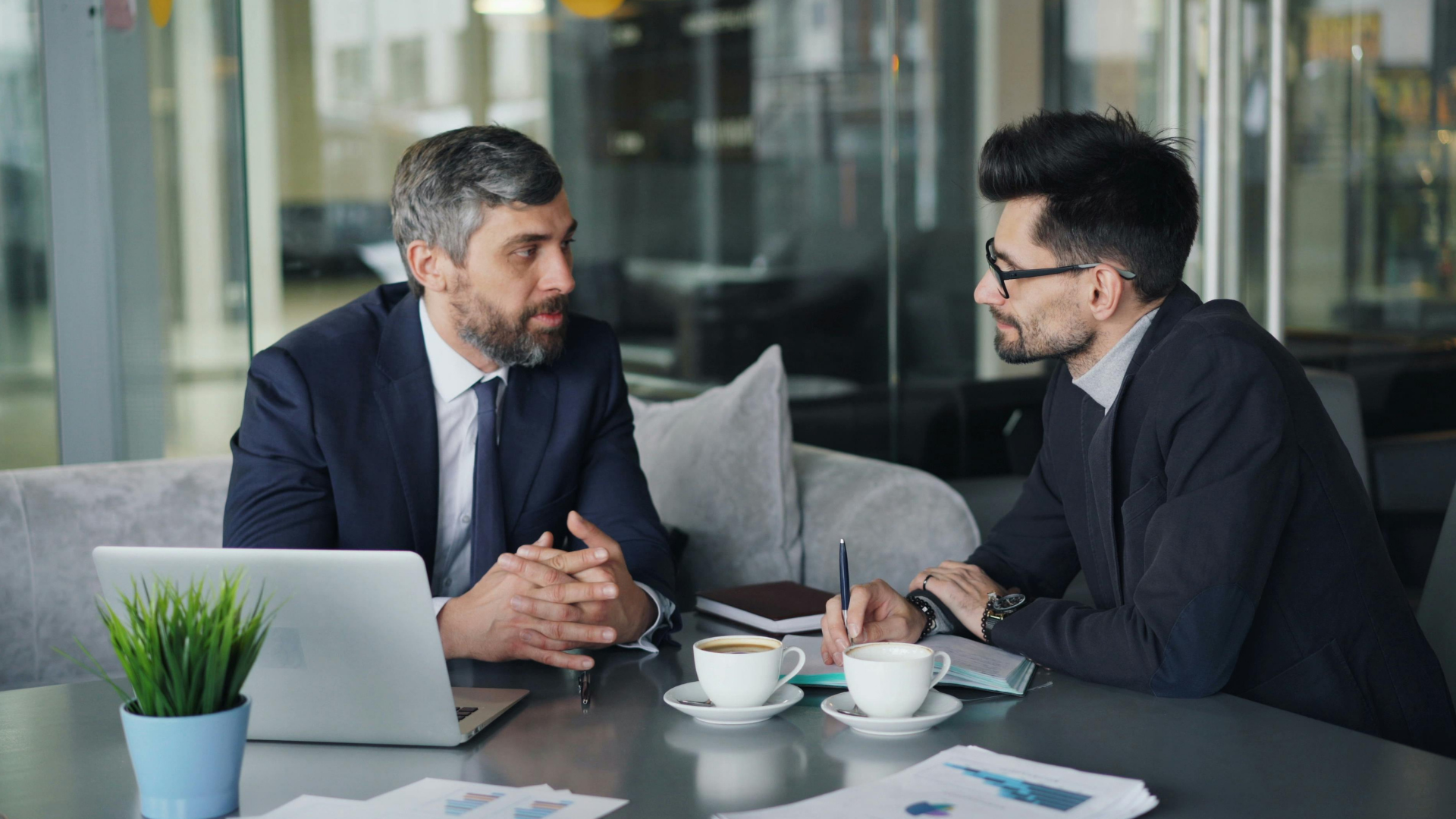 Two businesspeople in suits discussing at a table with a laptop, coffee cups, and papers in a modern office