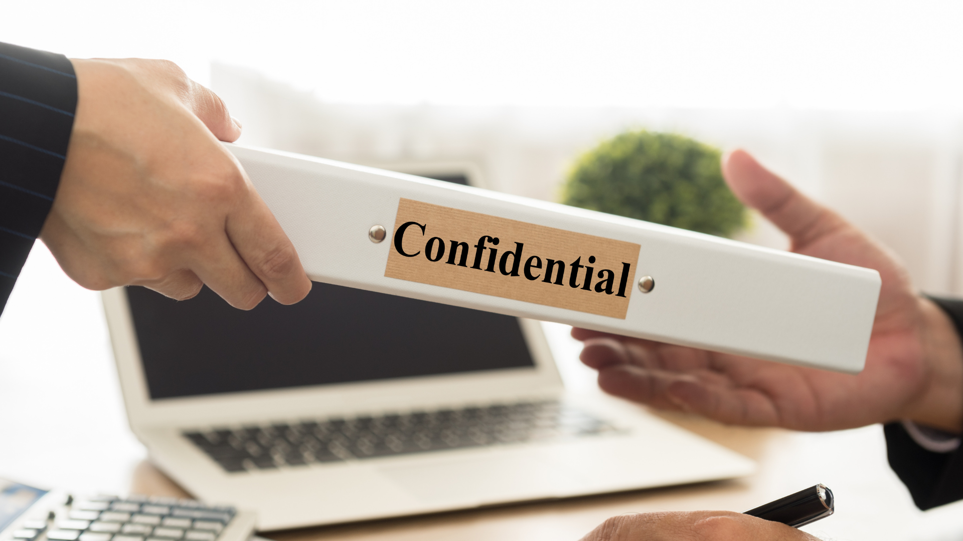 Hands passing a white folder labeled “Confidential” over a laptop on a desk