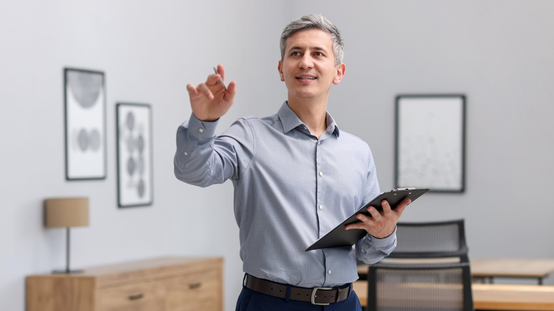 Man holding a tablet and pointing in a bright office with framed art and a desk lamp