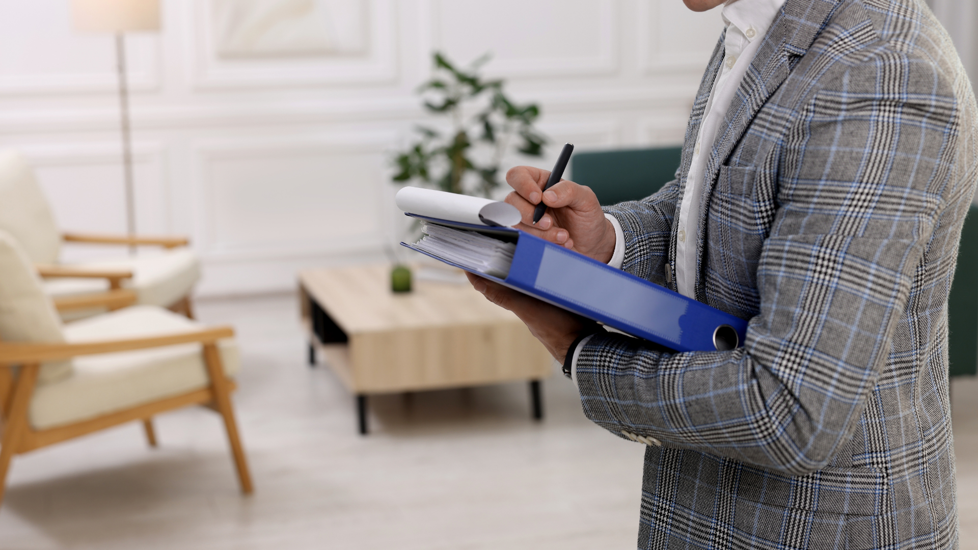 Person in a plaid suit writing on a clipboard inside a living room with chairs, a table, and a plant.