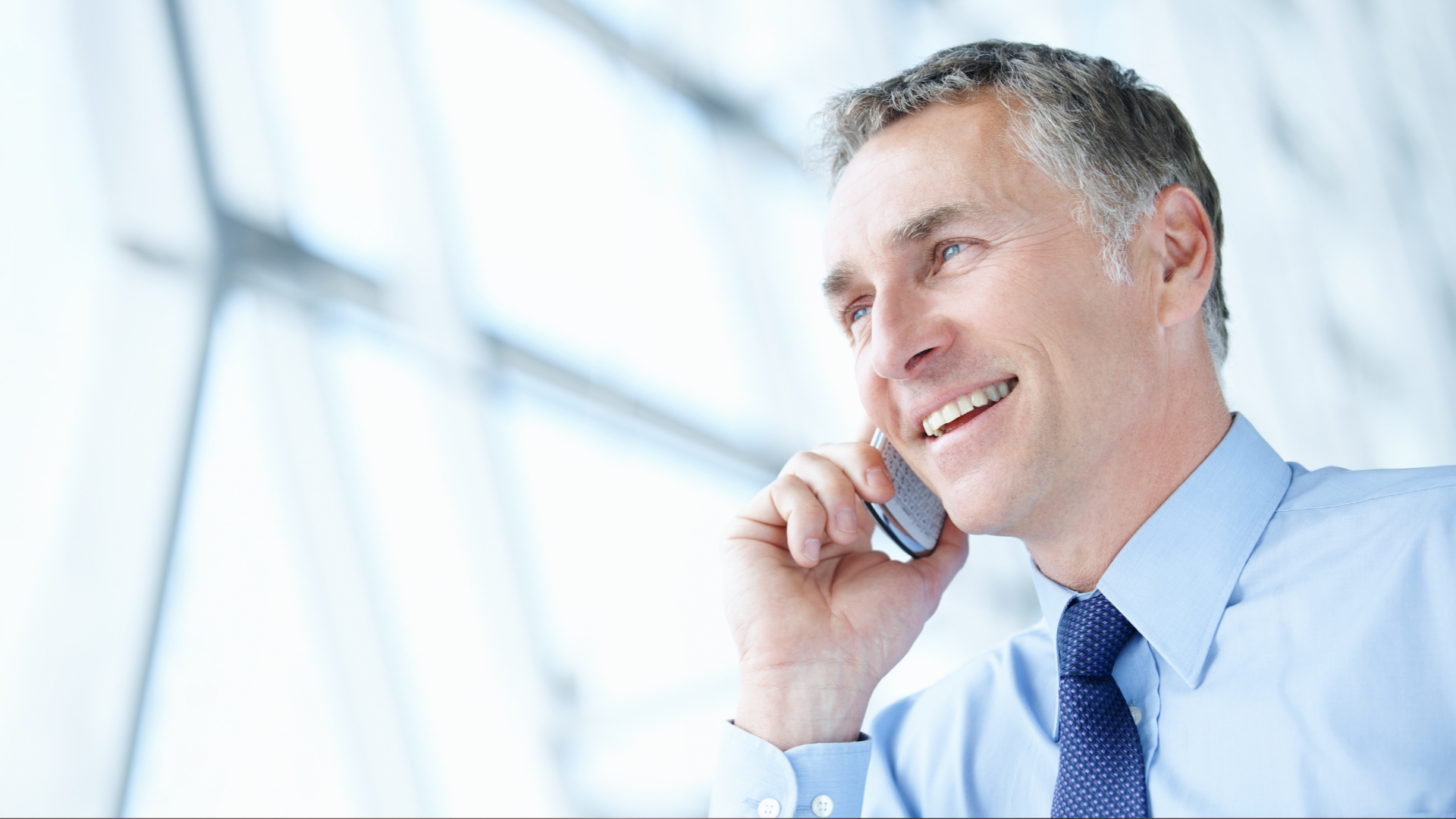 Smiling man in a blue shirt and tie talks on a phone in a bright office.