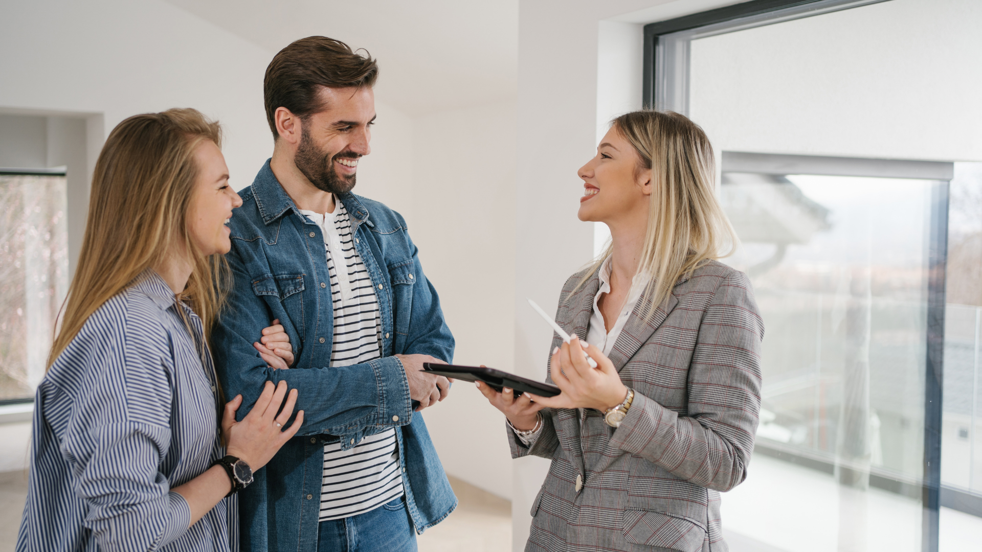 A real estate agent showing a couple a home; holding a tablet and pen, talking, smiles.