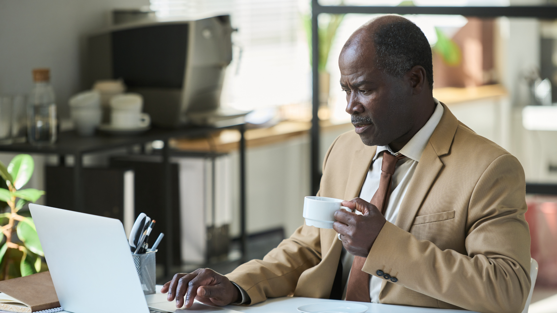 Man in tan suit, holding a cup, looking at a laptop in a well-lit office.
