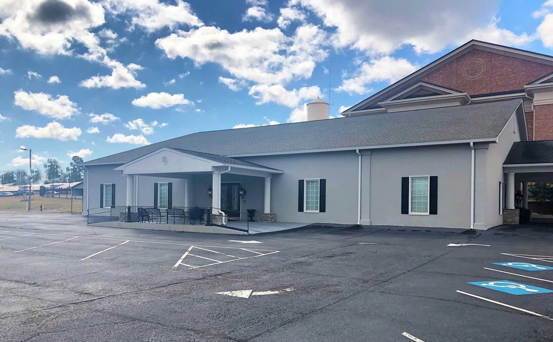 Gray building with dark shutters and a red-brick section, set on a parking lot with blue handicap parking symbols.