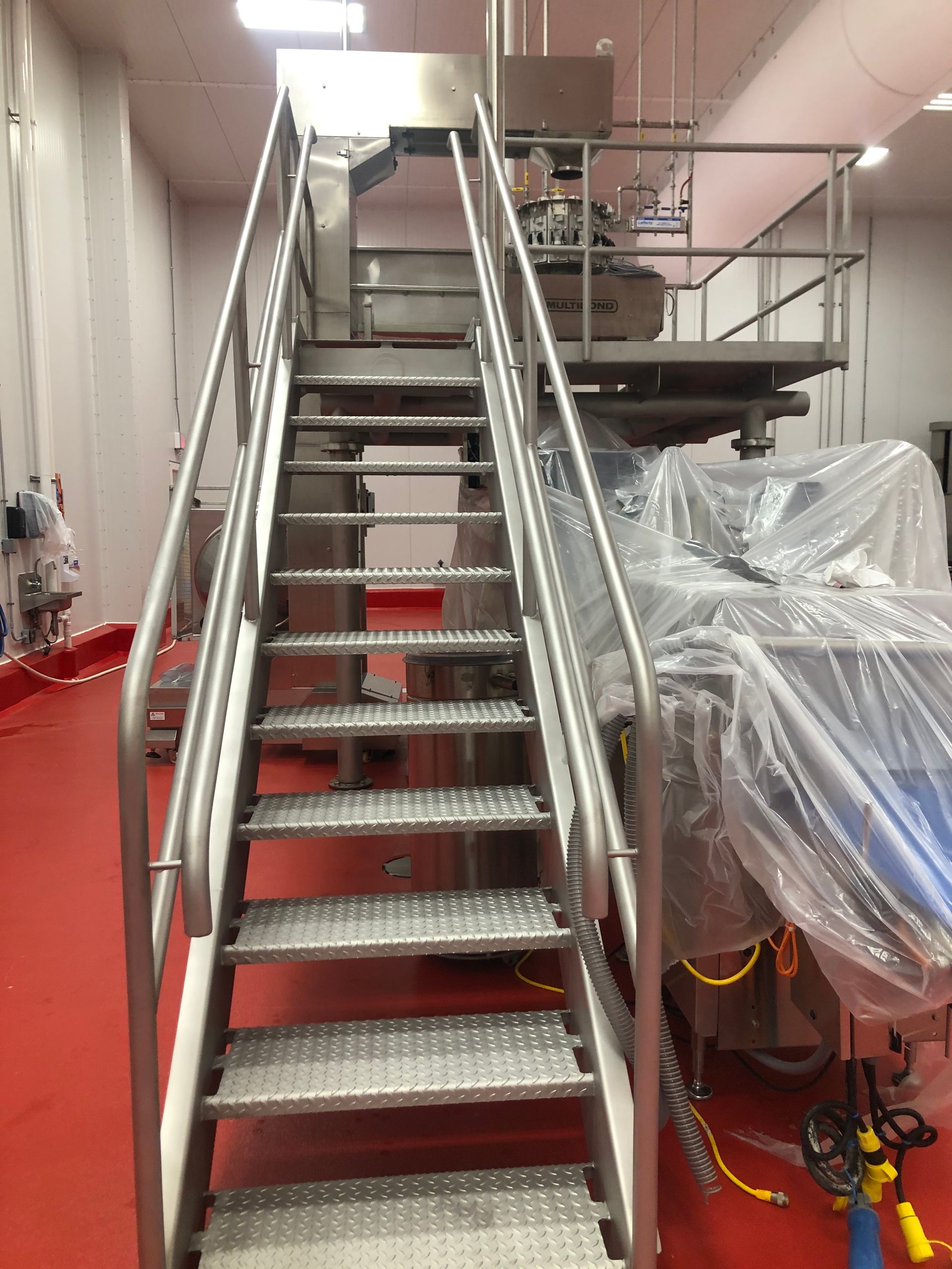 A metal staircase with textured treads leads to an elevated platform in a factory with a red floor.