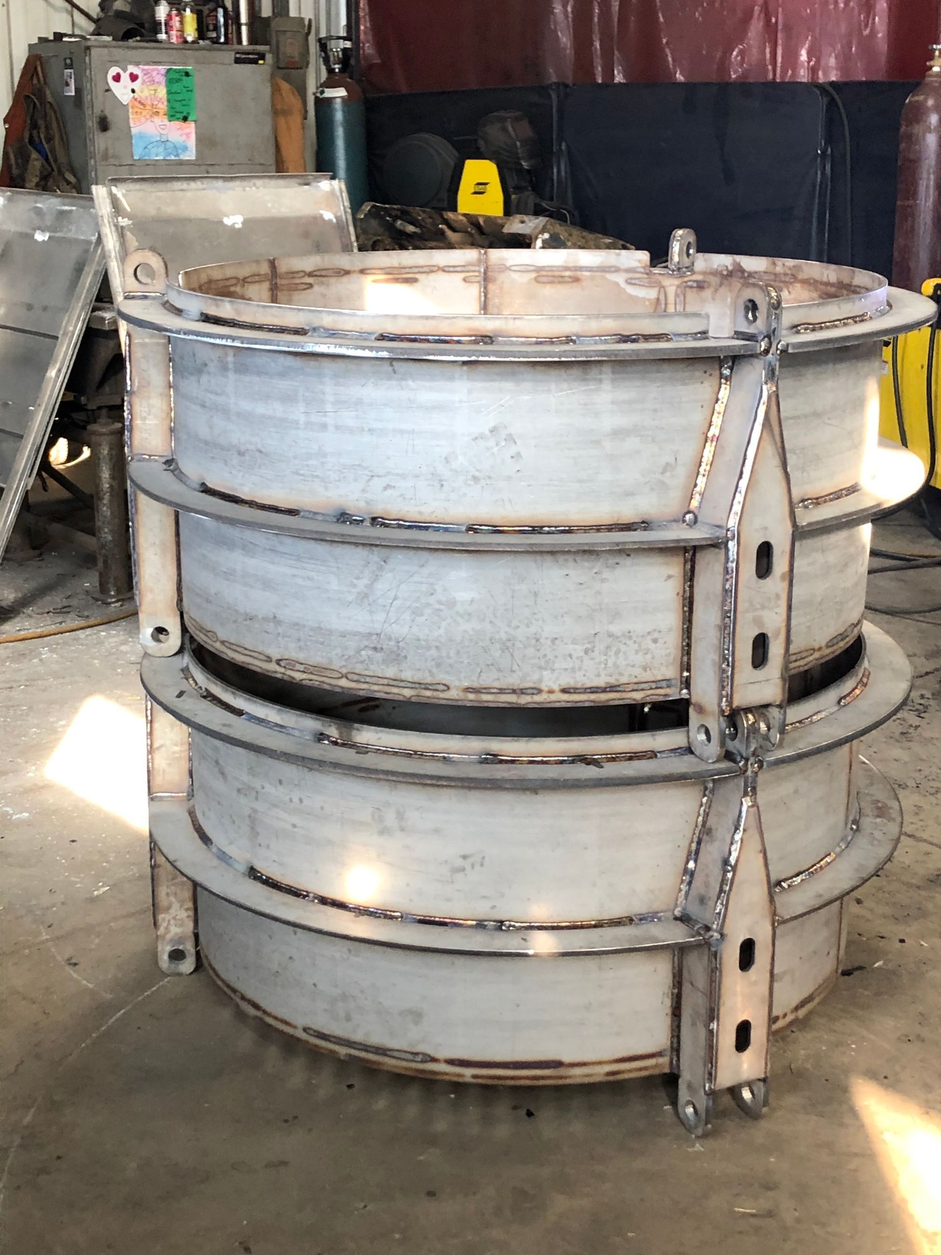 A custom fabricated metal cylindrical structure with welded steel rings and support beams in a workshop setting.
