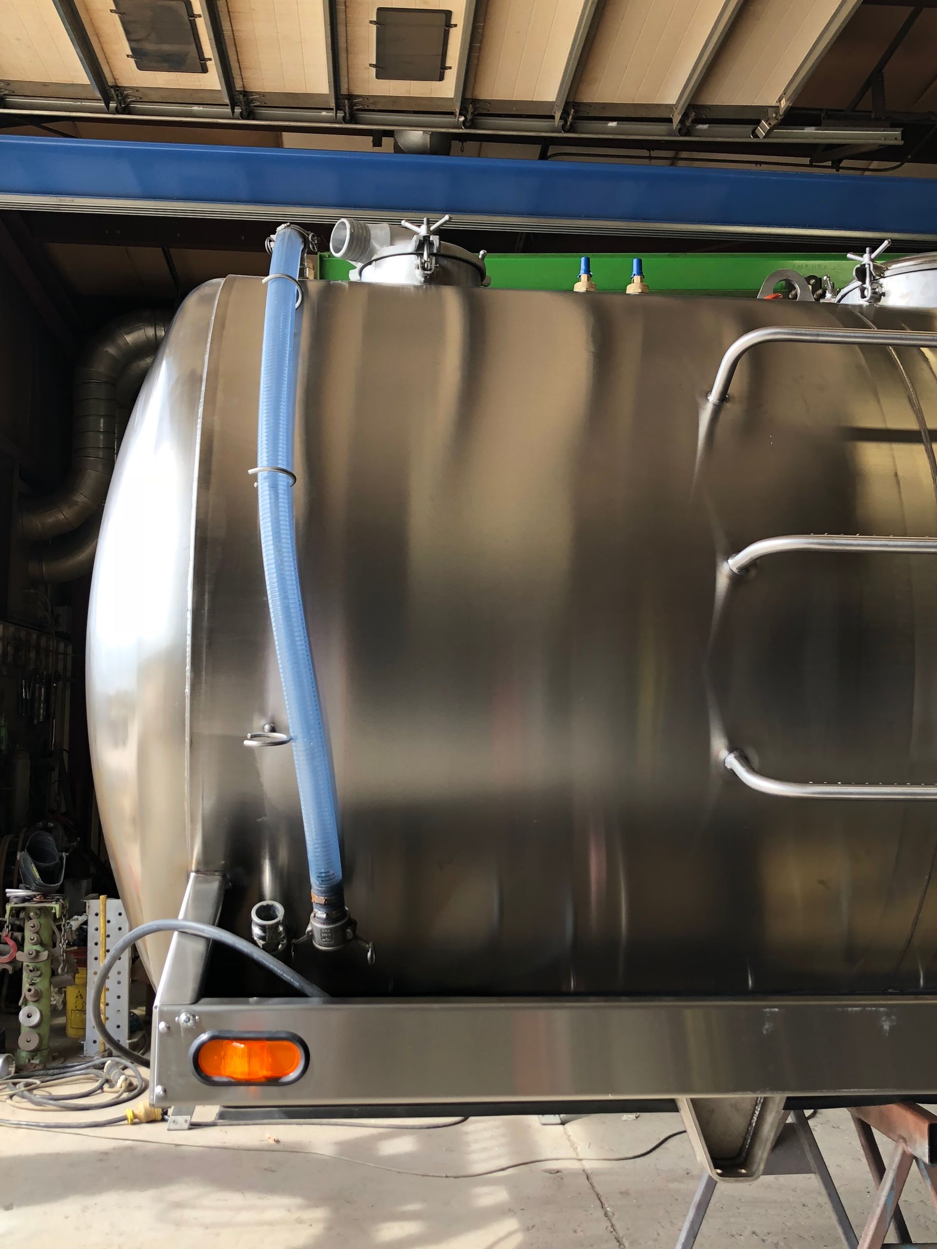 A stainless steel cylindrical tank trailer with an attached blue hose, mounted on a frame with an orange side light.