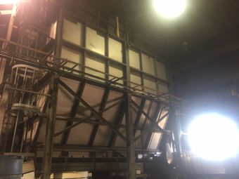 A large industrial metal hopper structure with a braced framework, seen from below in a dimly lit factory setting.