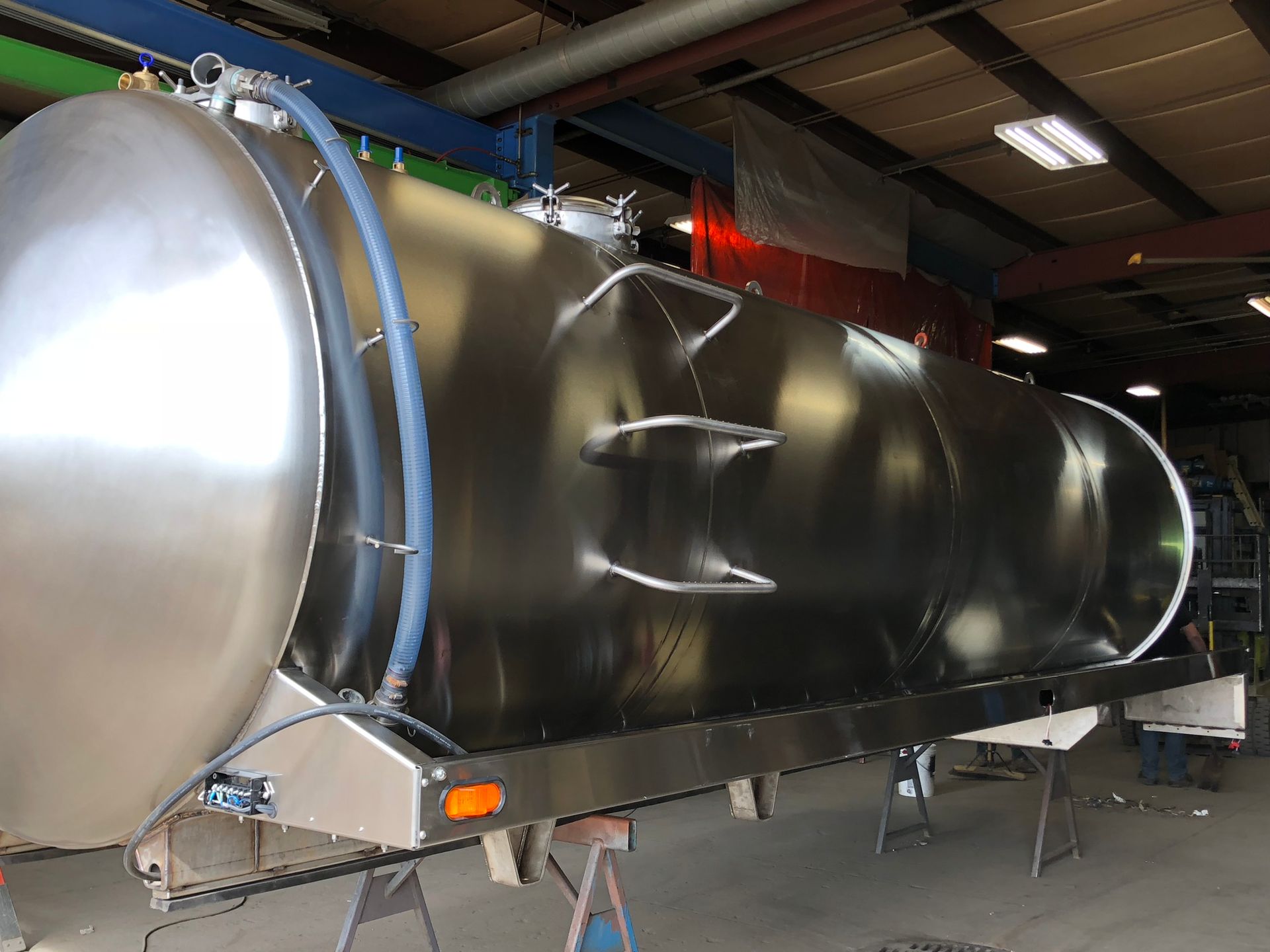 A large, polished stainless steel cylindrical tanker resting on metal supports inside a bright workshop.