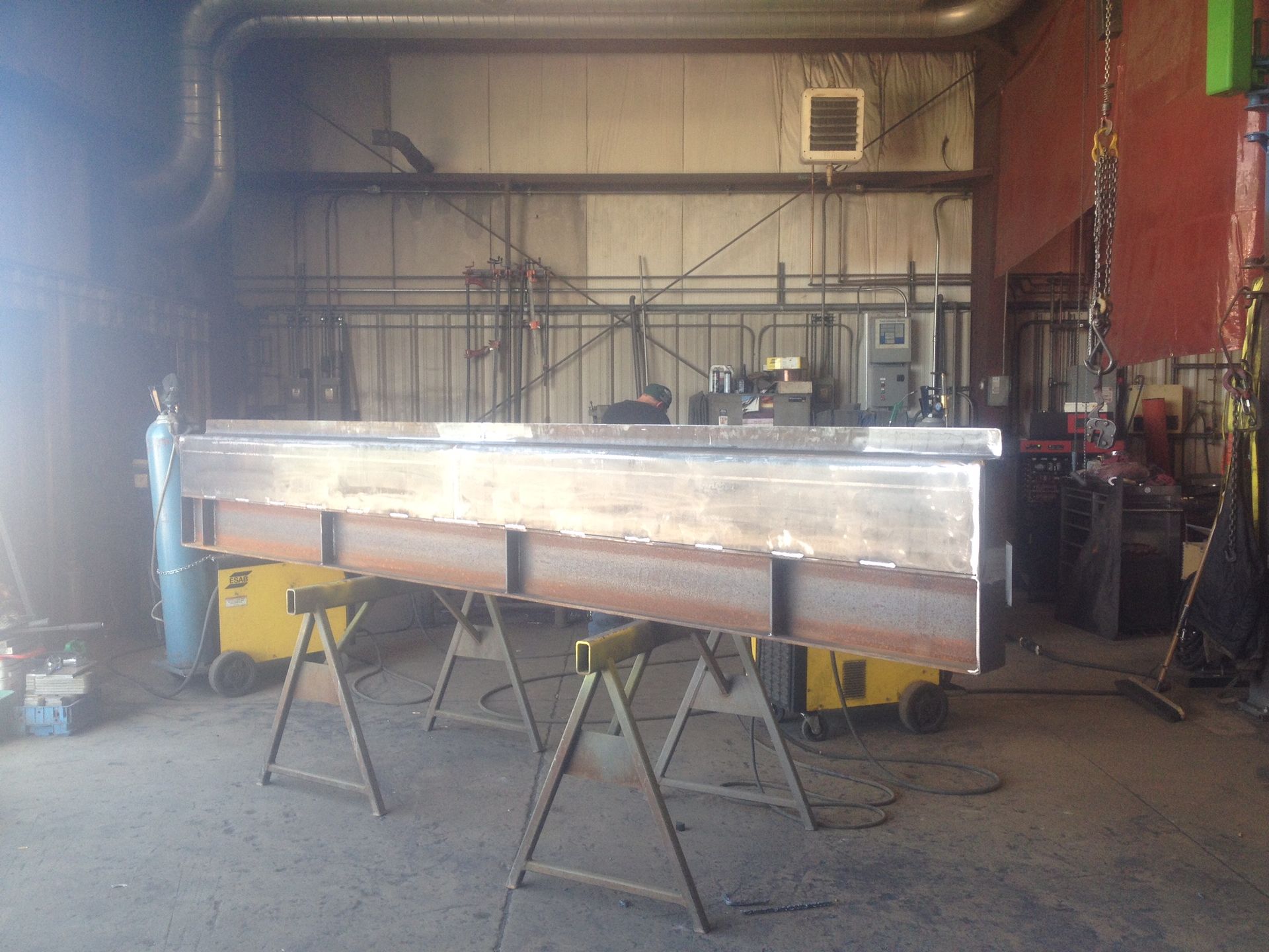 A long steel beam with a finished top section sits on metal sawhorses inside a welding workshop.