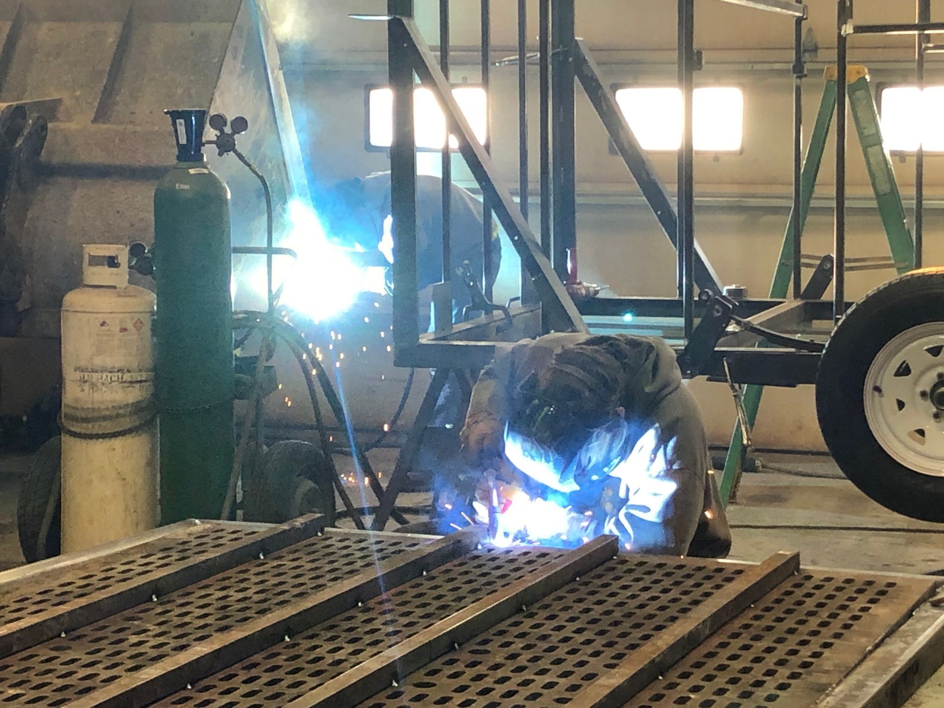 A welder wearing a protective mask works on a metal frame inside a workshop with a gas tank and a ladder nearby.