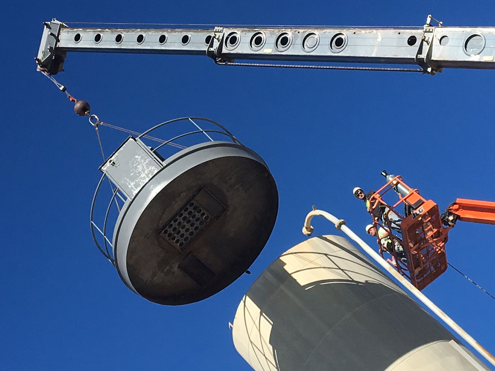 A crane lowers a large circular structure with a railing onto a vertical tank as workers in a basket assist nearby.