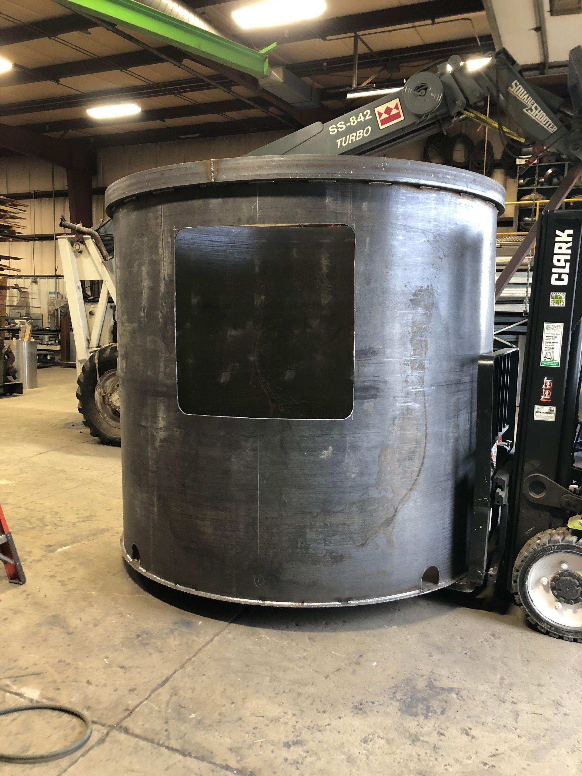 A large, dark metal cylinder with a square dark panel attached, being moved by a forklift inside a workshop.