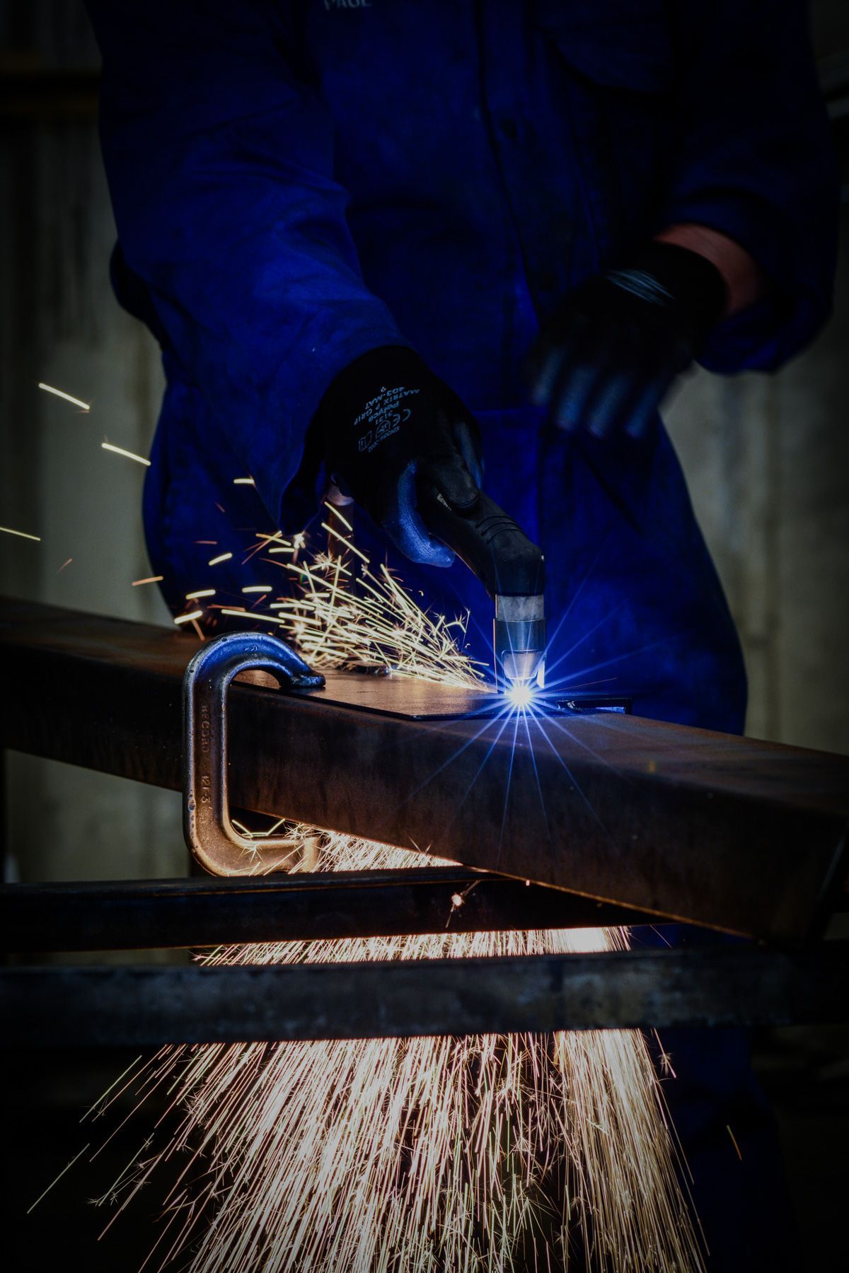 A person wearing blue workwear and gloves uses a plasma cutter to slice through a metal beam, creating bright sparks.