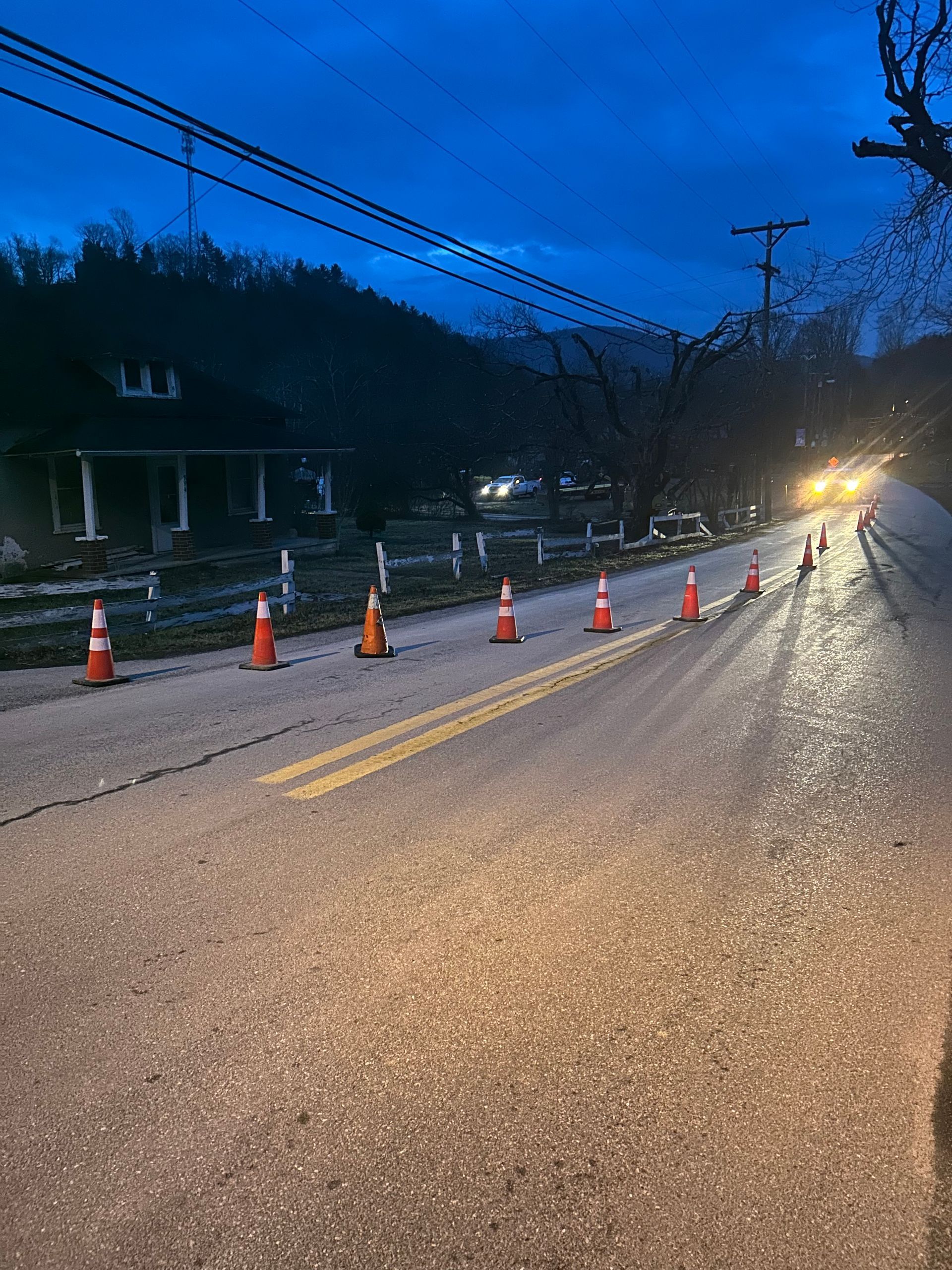 Dusk road with orange traffic cones blocking one lane, white barrier tape, and headlights approaching from the right