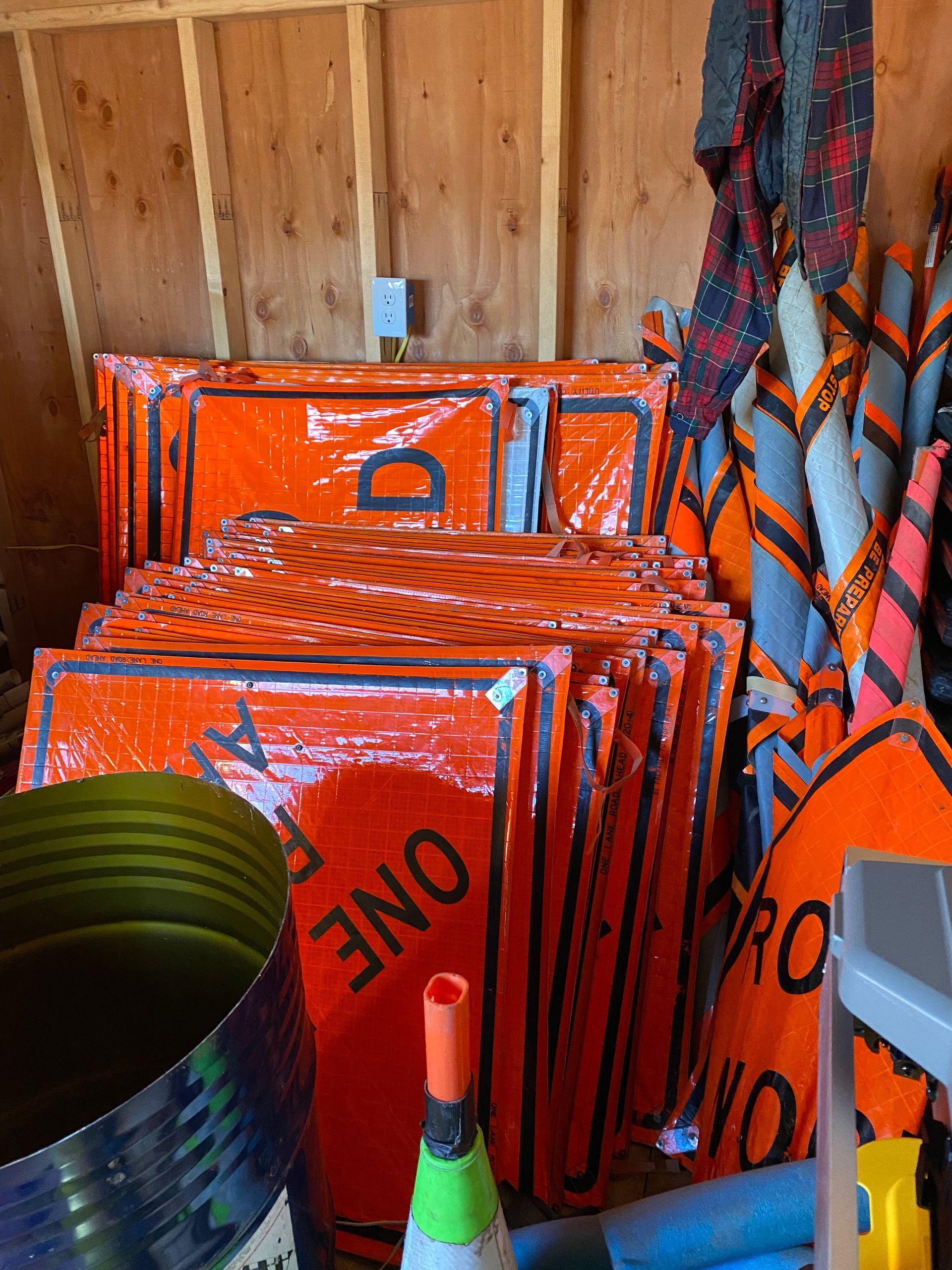 Stacked orange one-way signs and umbrellas in a wooden shed, with a striped bucket in the foreground.