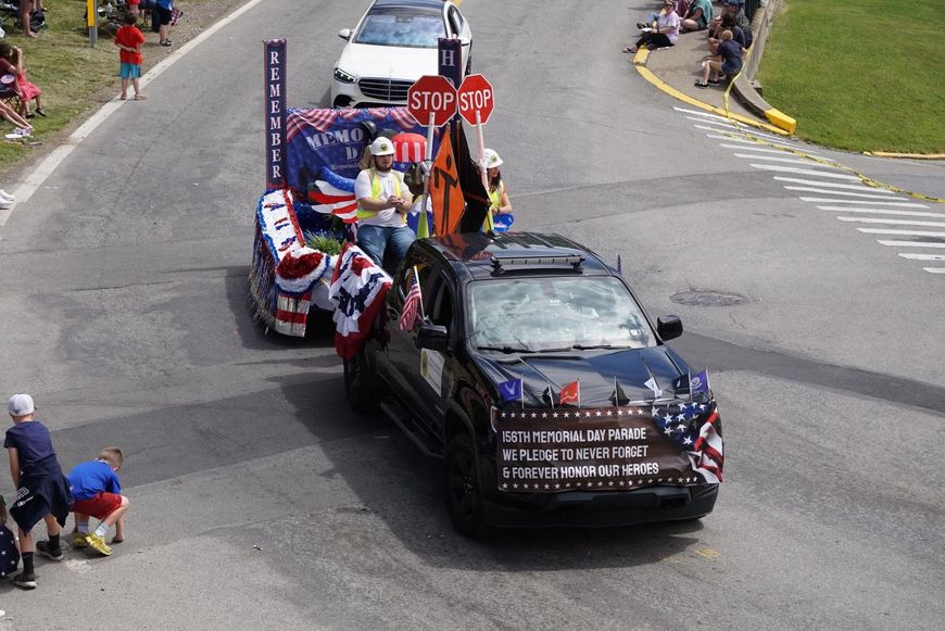 Decorated parade cars on a street corner with a STOP sign and spectators watching nearby