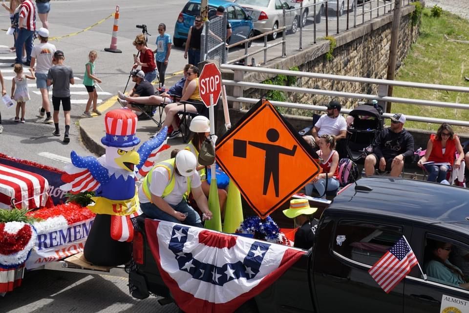 Parade float with patriotic bunting, people, and a large orange roadwork sign on a city street