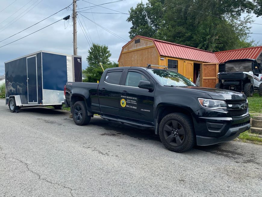 Black pickup truck parked on a street beside a blue enclosed trailer and a small wooden building