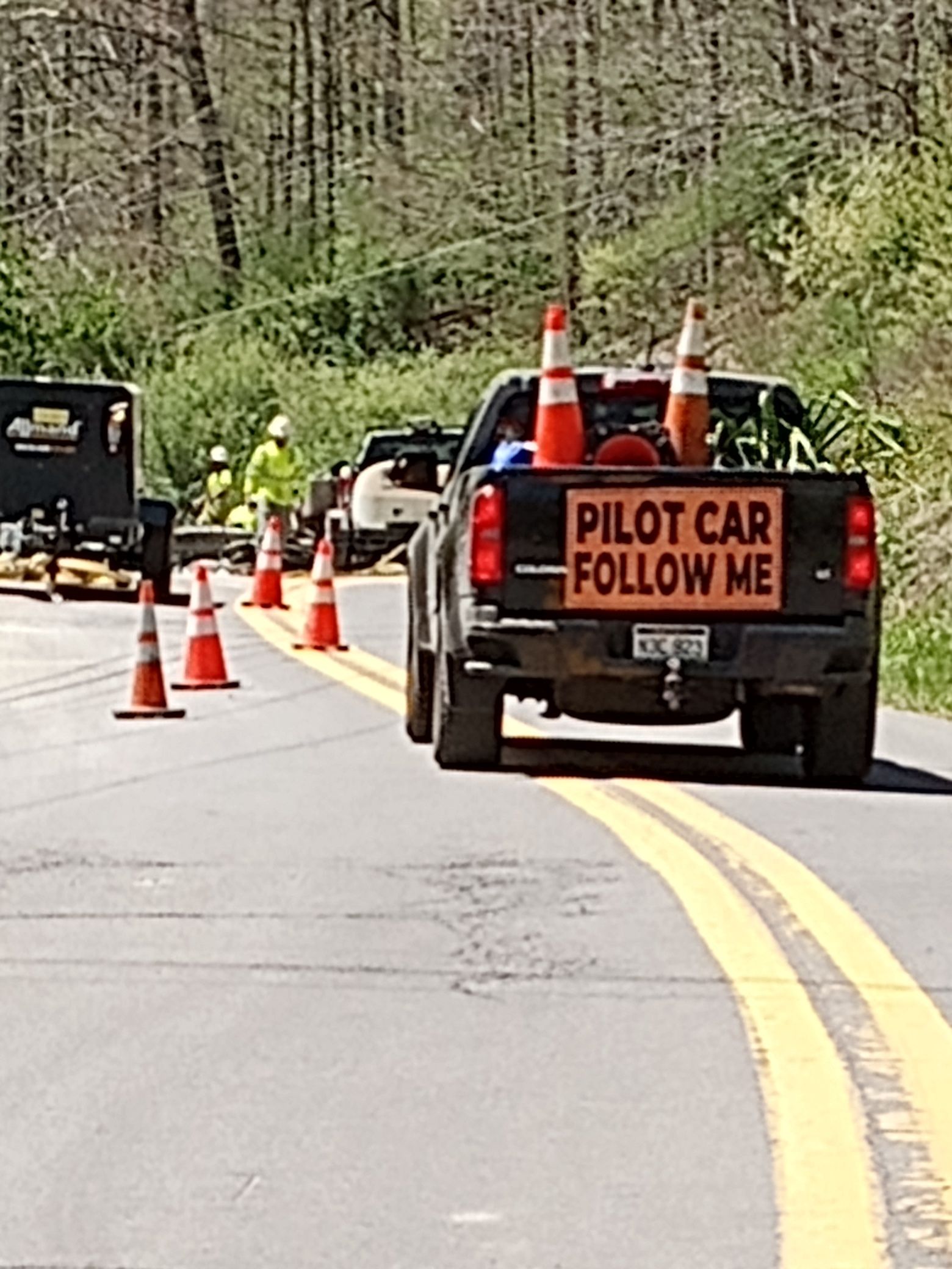 Roadwork scene with a pickup truck marked “PILOT CAR FOLLOW ME” and orange cones on a curving road