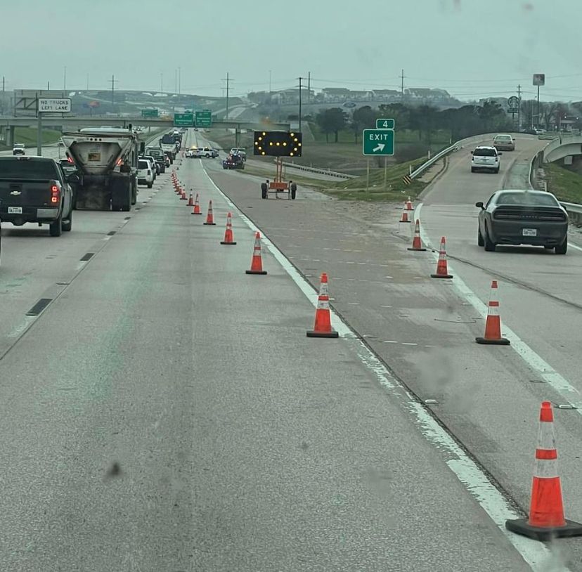 Traffic cones lane closure on a highway with vehicles driving past under overcast skies