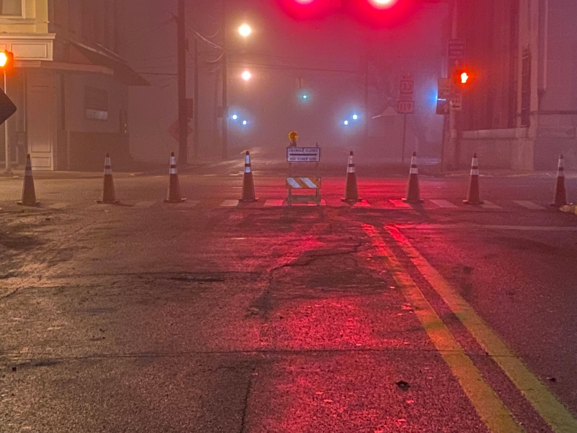 Foggy city street at night with red traffic lights, wet pavement, and traffic cones blocking the road