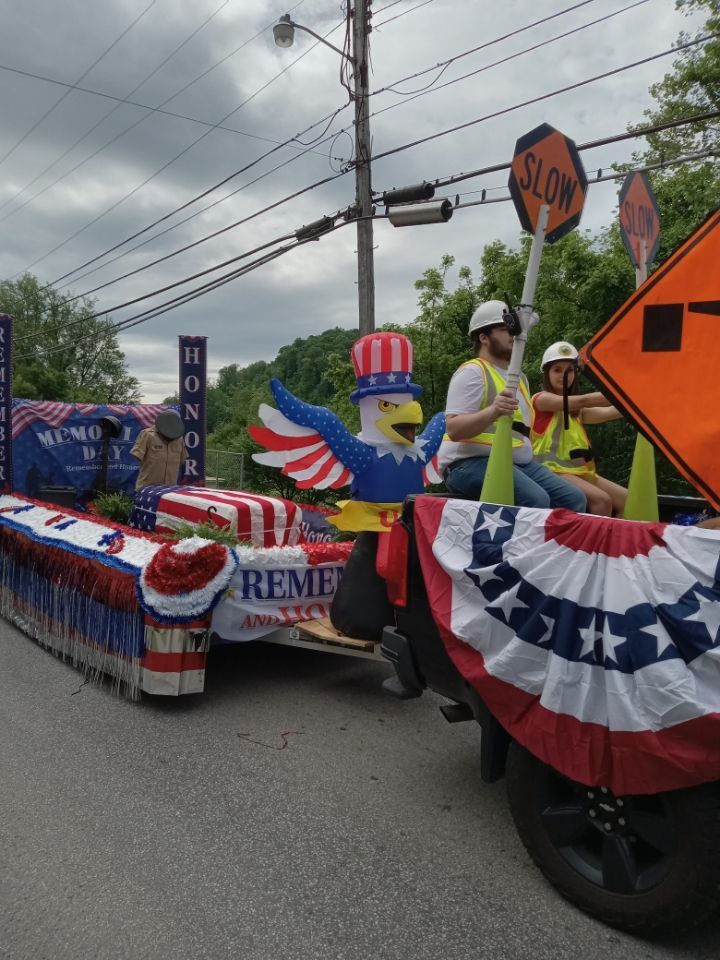 Patriotic parade float with red, white, and blue bunting and flags, moving past a stop sign under cloudy skies