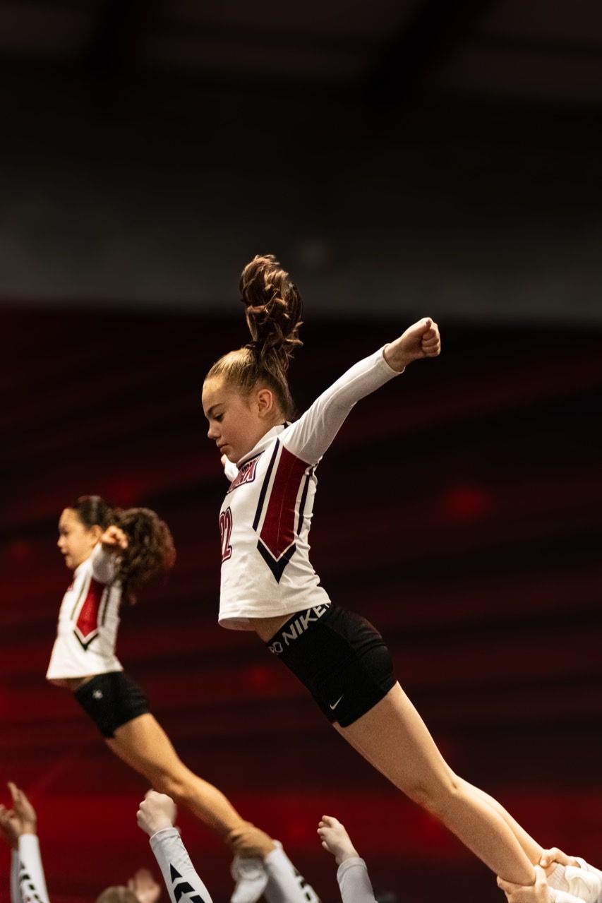 Two cheerleaders in maroon and white uniforms leap during a routine on a dim stage