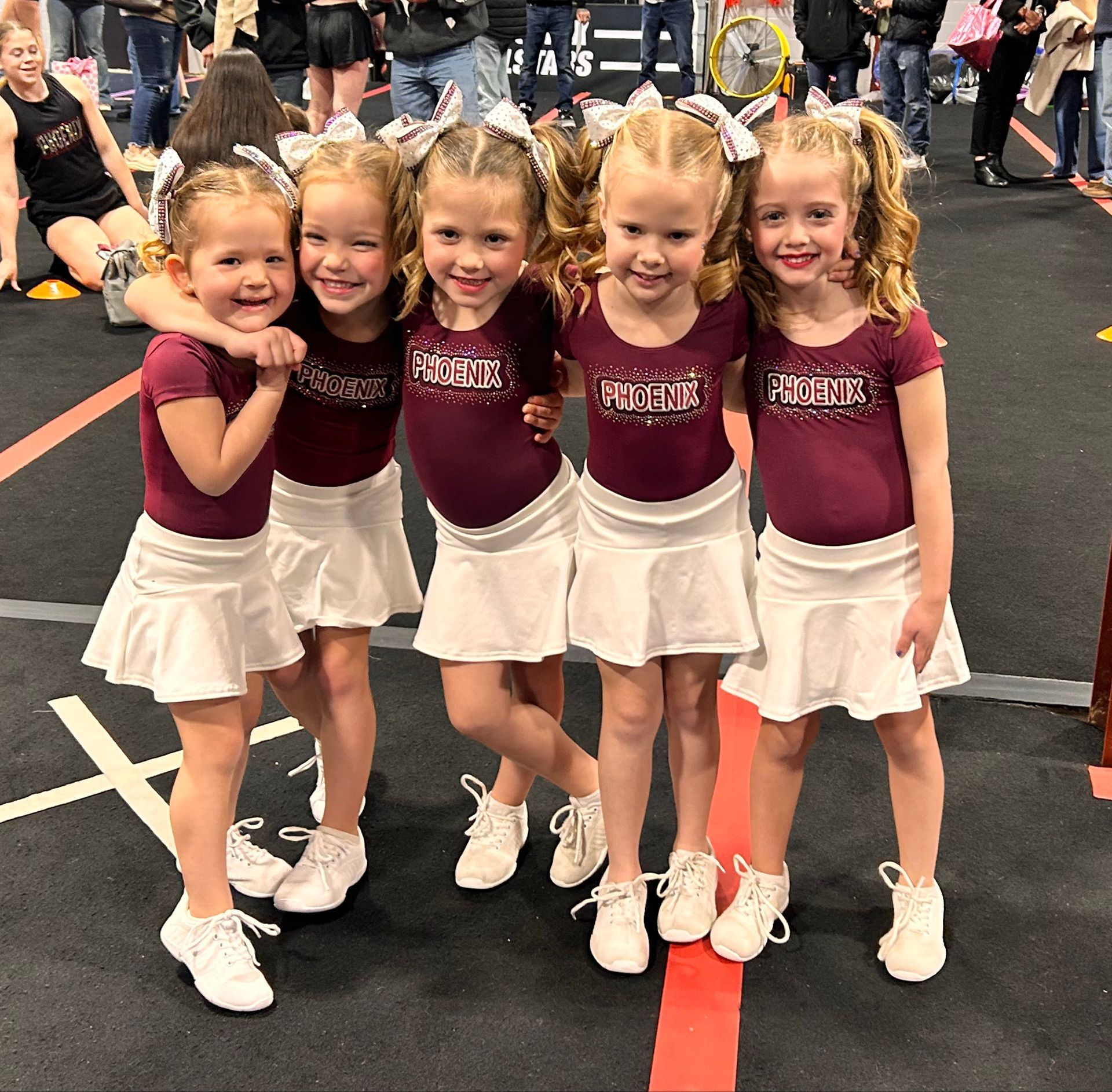 Five young cheerleaders in maroon tops and white skirts posing together indoors at a competition