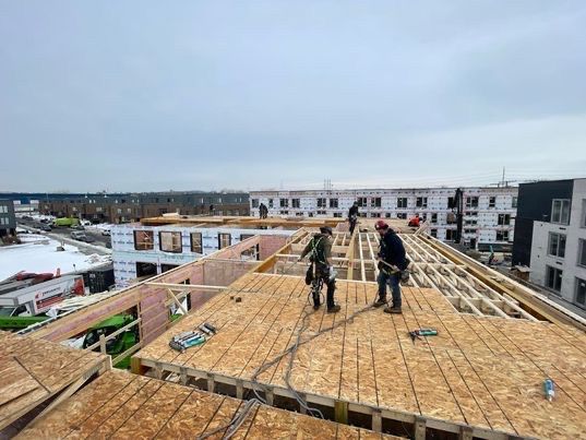 Des ouvriers du bâtiment, outils à la main, installent des plaques de bois sur un toit, sous un ciel couvert.