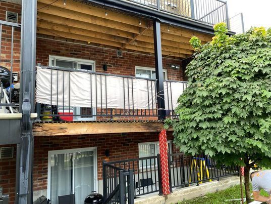 Immeuble en briques de plusieurs étages avec balcons, balustrades noires et brise-vues blancs. Un arbre se trouve à droite.