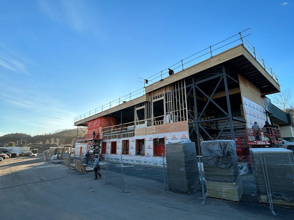 Chantier : charpente apparente, murs inachevés et piles de bois. Ouvriers visibles. Ciel bleu.