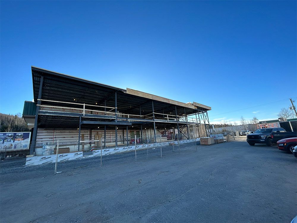 Chantier de construction d'un bâtiment de deux étages avec structure métallique apparente, parking en gravier, ciel bleu dégagé.