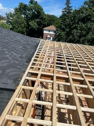 Toiture en construction ; poutres en bois et charpente apparente ; toit en bardeaux gris foncé à gauche, ciel bleu, arbres en arrière-plan.
