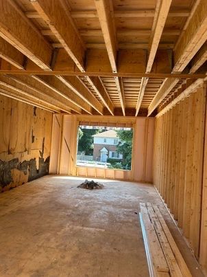 Vue intérieure d'un bâtiment en construction, montrant la charpente apparente en bois. Une fenêtre donne sur une maison.