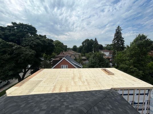 Vue d'un toit plat recouvert de contreplaqué, d'arbres et de maisons sous un ciel nuageux.