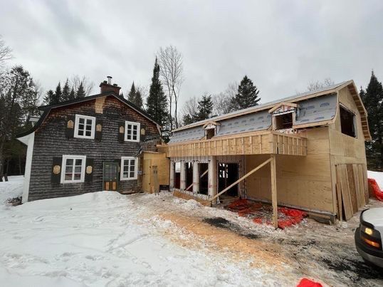 Rénovation d'une maison dans un cadre enneigé : une vieille cabane en rondins jouxte une nouvelle structure en bois en construction.