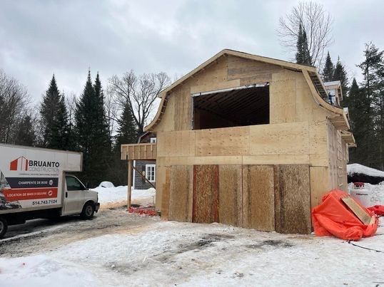 Construction d'un bâtiment en bois de deux étages avec un toit incurvé. Un camion est stationné à proximité et le sol est recouvert de neige.