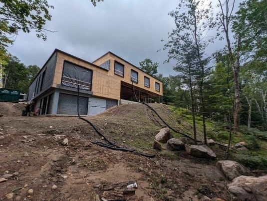 Maison moderne de deux étages en construction sur un terrain boisé en pente. Les murs extérieurs ne sont pas terminés.