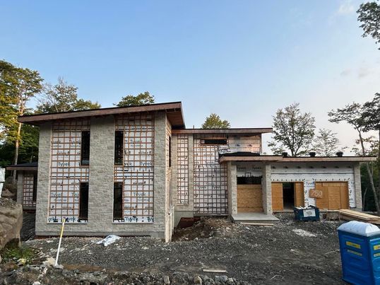 Maison neuve en construction, vue extérieure. Murs en briques grises, porte de garage en bois, ciel bleu.